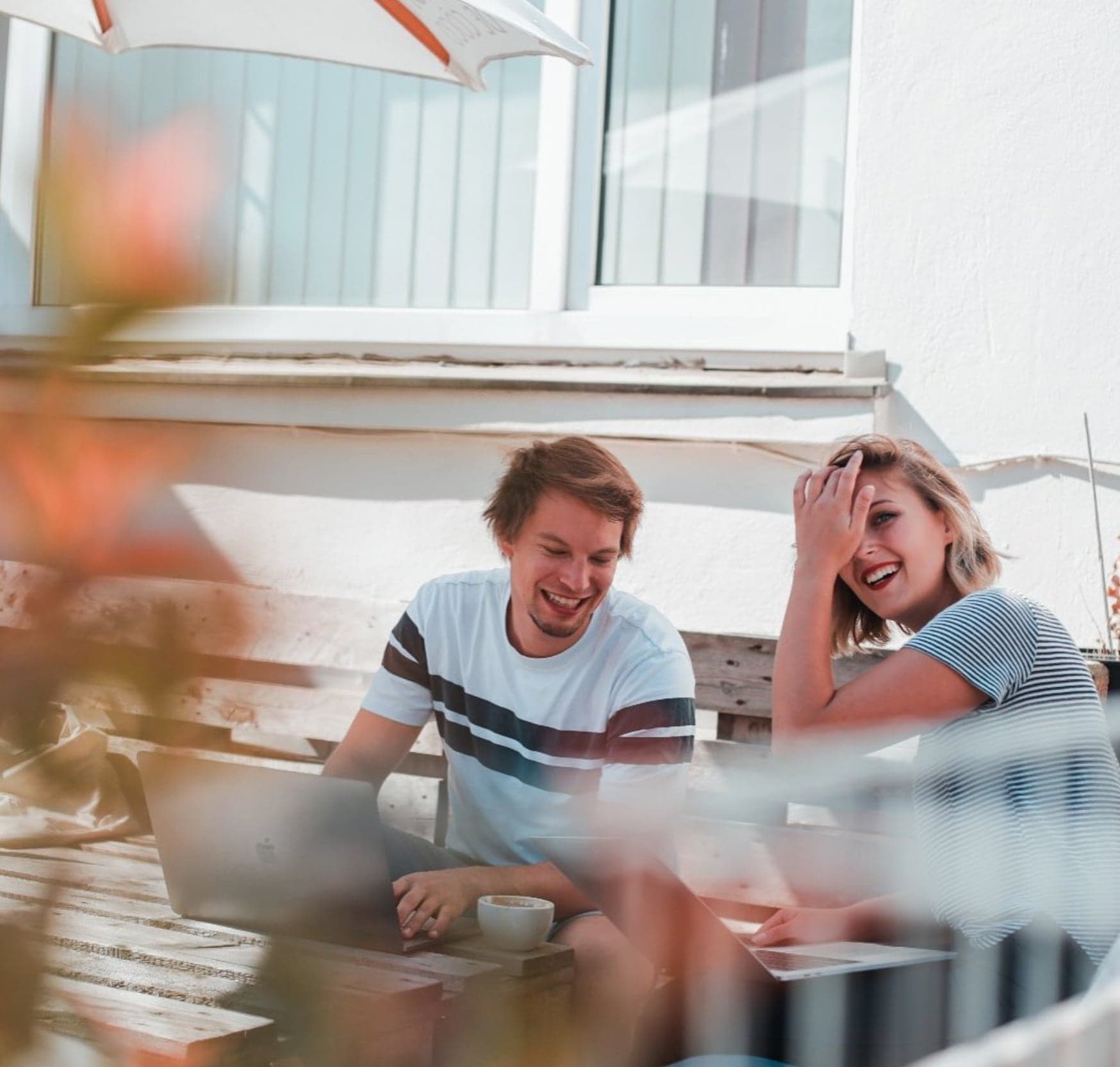 Corina Renner und ein Kollege lachend beim gemeinsamen Arbeiten am Laptop auf der Terrasse